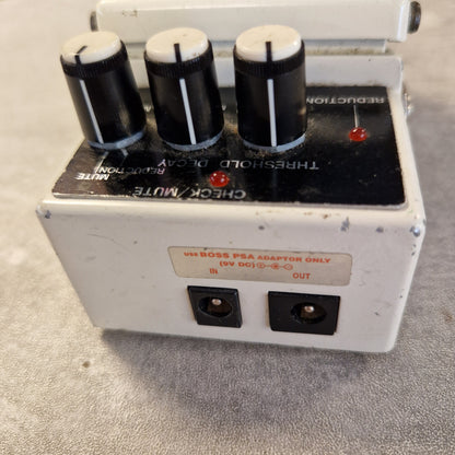Pre-Owned Boss NS-2 Noise Suppressor Pedal A vintage-style guitar effects pedal with three black knobs, red indicator lights, and a white metal body on a gray textured surface. The front label reads 'CHECK / MUTE' and 'THRESHOLD DECAY' with red text.