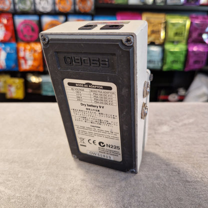 Pre-Owned Boss NS-2 Noise Suppressor Pedal A BOSS AC Adapter power supply unit with a white casing and a black front faceplate, showing the model number 19093 and a 9V battery label. The unit is placed on a gray textured surface with a blurred background of colorful retail items.