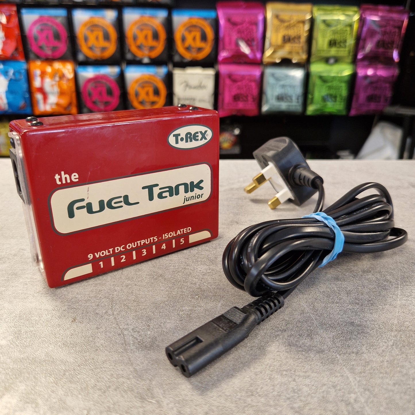Pre-Owned T. Rex Fuel Tank Junior Power Supply A red T-REX 'The Fuel Tank Junior' 9V DC output pedal shown next to its black UK plug cable on a gray tabletop, with guitar effects pedals in the background.