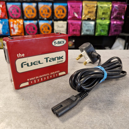 Pre-Owned T. Rex Fuel Tank Junior Power Supply A red T-REX 'The Fuel Tank Junior' 9V DC output pedal shown next to its black UK plug cable on a gray tabletop, with guitar effects pedals in the background.