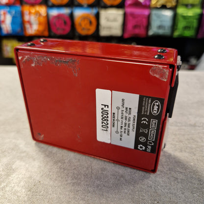 Pre-Owned T. Rex Fuel Tank Junior Power Supply A red rectangular electronic module with a white and black label, photographed on a gray surface with blurred colorful items in the background.