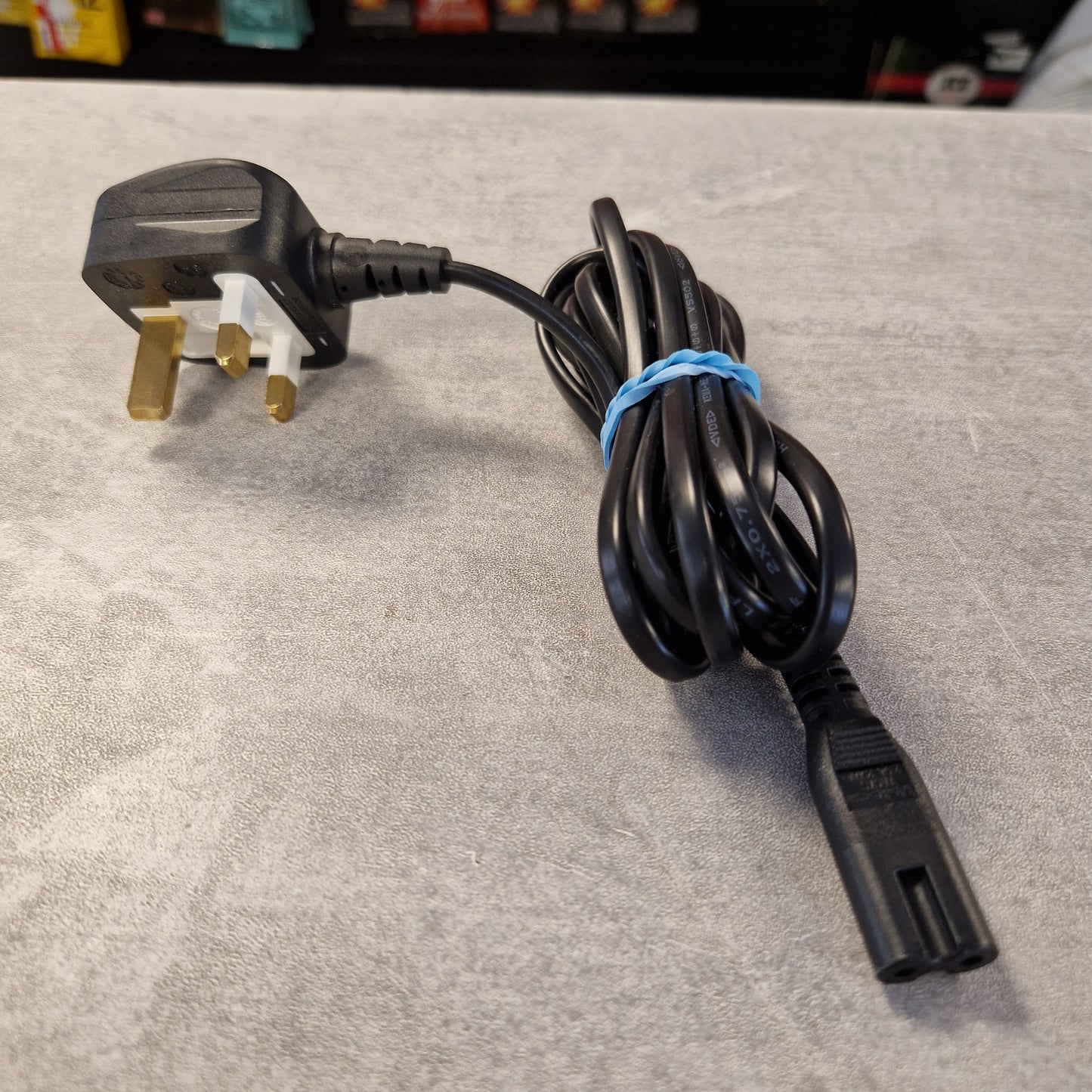 Pre-Owned T. Rex Fuel Tank Junior Power Supply A black three-pin UK wall plug power cord with a coiled black cable secured by a blue rubber band, resting on a gray textured surface.