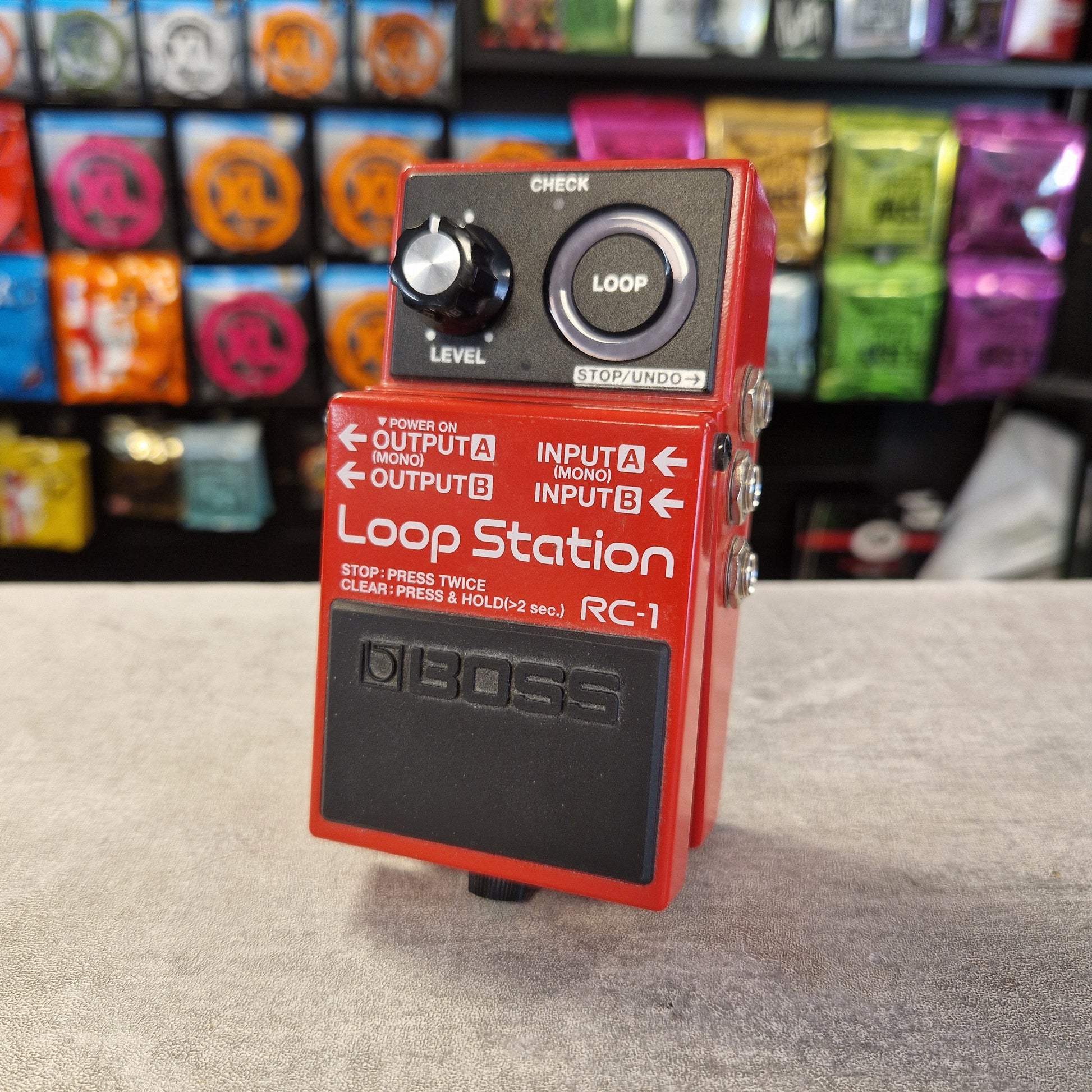 Pre-Owned Boss RC-1 Loop Station Pedal A red and black Boss RC-1 Loop Station guitar effects pedal standing upright on a gray surface, with a blurred shop background of colorful pedal cases.