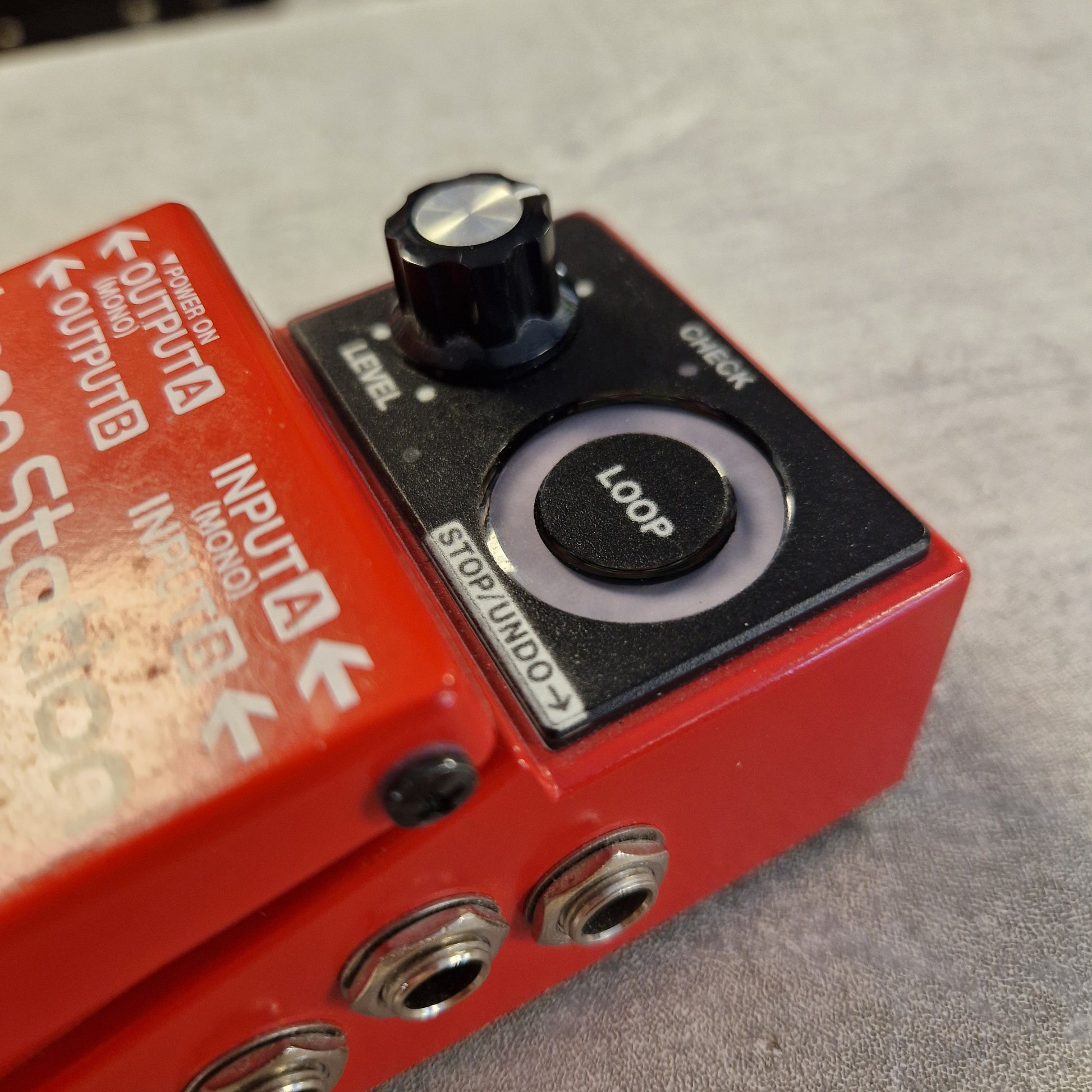 Pre-Owned Boss RC-1 Loop Station Pedal A close-up of a red and black Uni-Music Studio 1000 guitar effects pedal with a black loop knob and input/output jacks, resting on a gray textured surface.