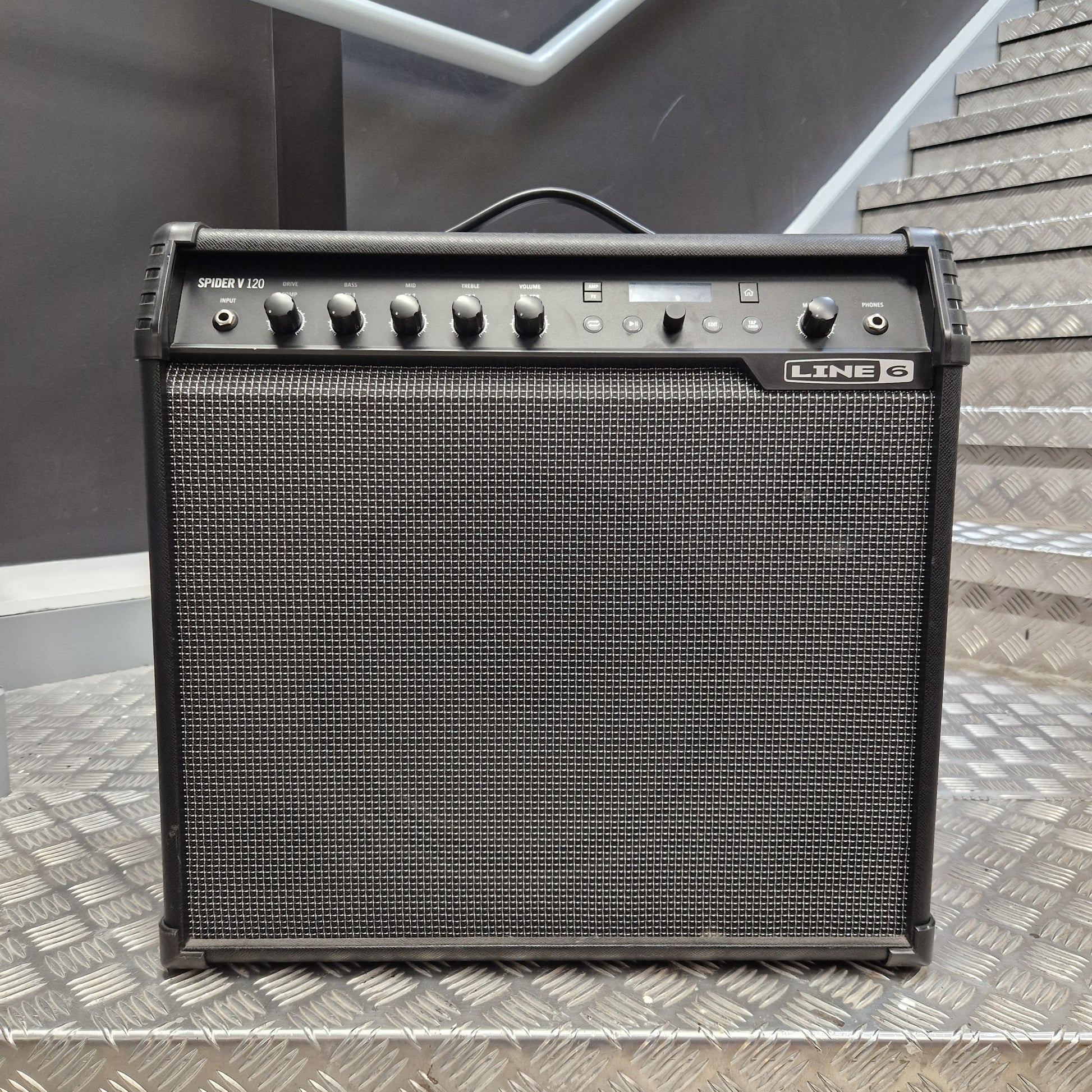 Pre-Owned Line 6 Spider V 120 Mk II Guitar Combo Amplifier A black Line 6 Synergy 700 guitar amplifier with a front speaker grille, set on a diamond plate metal surface with stairs in the background.