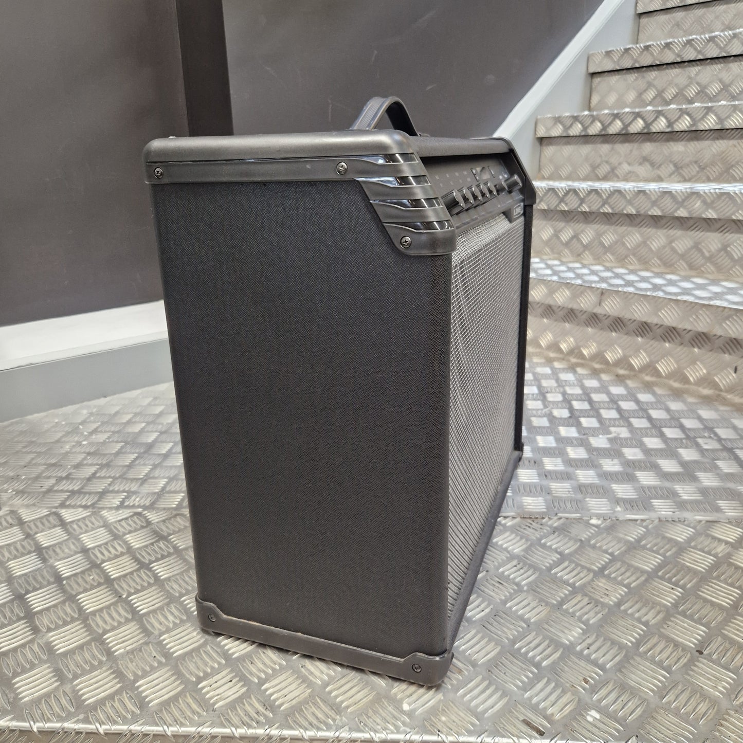 Pre-Owned Line 6 Spider V 120 Mk II Guitar Combo Amplifier A black guitar amplifier with a textured fabric cover and a top handle, positioned on a diamond plate metal staircase.