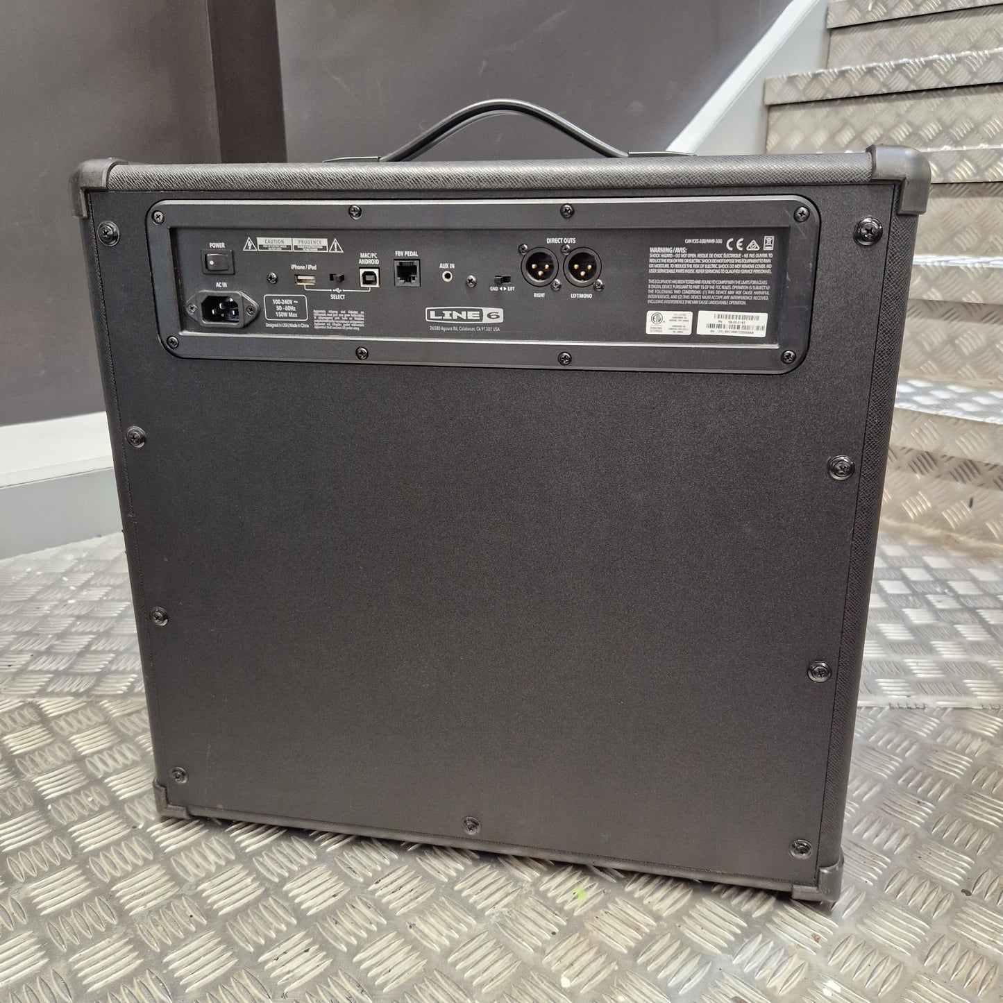 Pre-Owned Line 6 Spider V 120 Mk II Guitar Combo Amplifier A black guitar amplifier or speaker cabinet photographed from the back, standing on a diamond-plate metal floor with stairs in the background.