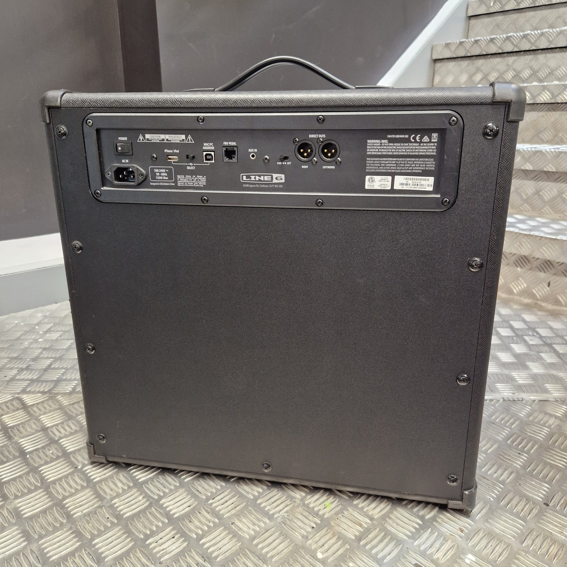 Pre-Owned Line 6 Spider V 120 Mk II Guitar Combo Amplifier A black guitar amplifier or speaker cabinet photographed from the back, standing on a diamond-plate metal floor with stairs in the background.