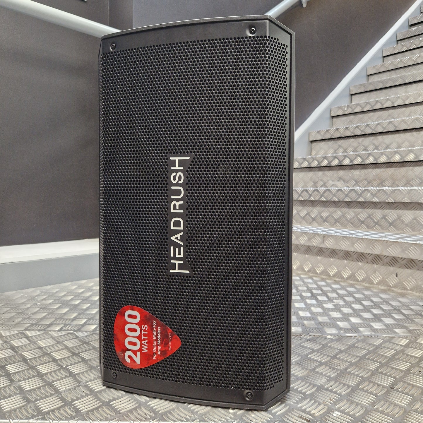 Pre-Owned Headrush FRFR-112 Full Range Powered 1x12 Speaker A black Headrush portable speaker with a red '2000' badge, standing on a diamond plate staircase landing.