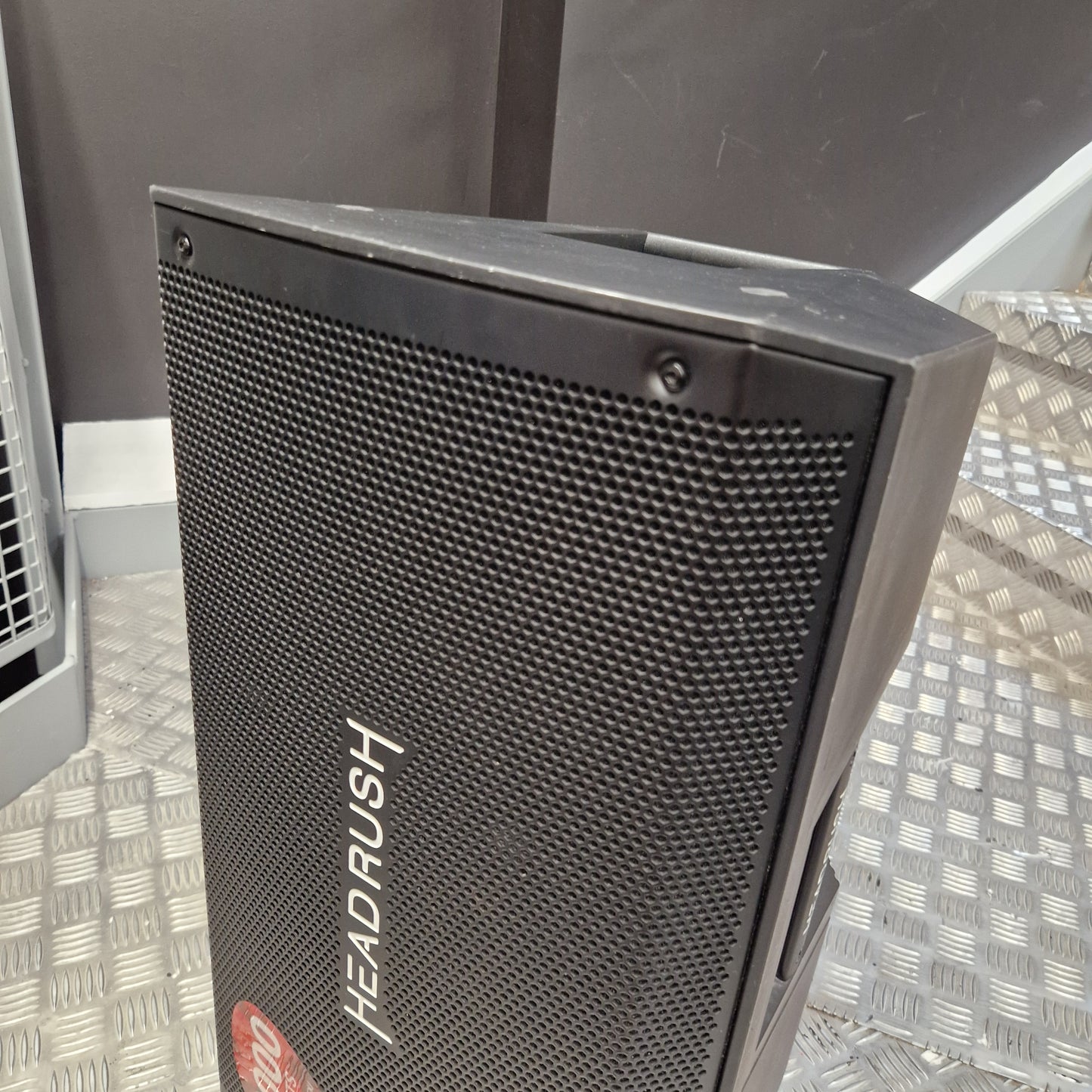 Pre-Owned Headrush FRFR-112 Full Range Powered 1x12 Speaker A black and gray portable speaker with a metal mesh grille, featuring the brand name 'HEAD RUSH' printed vertically on the front. The speaker is photographed on a diamond plate metal floor against a dark gray background.