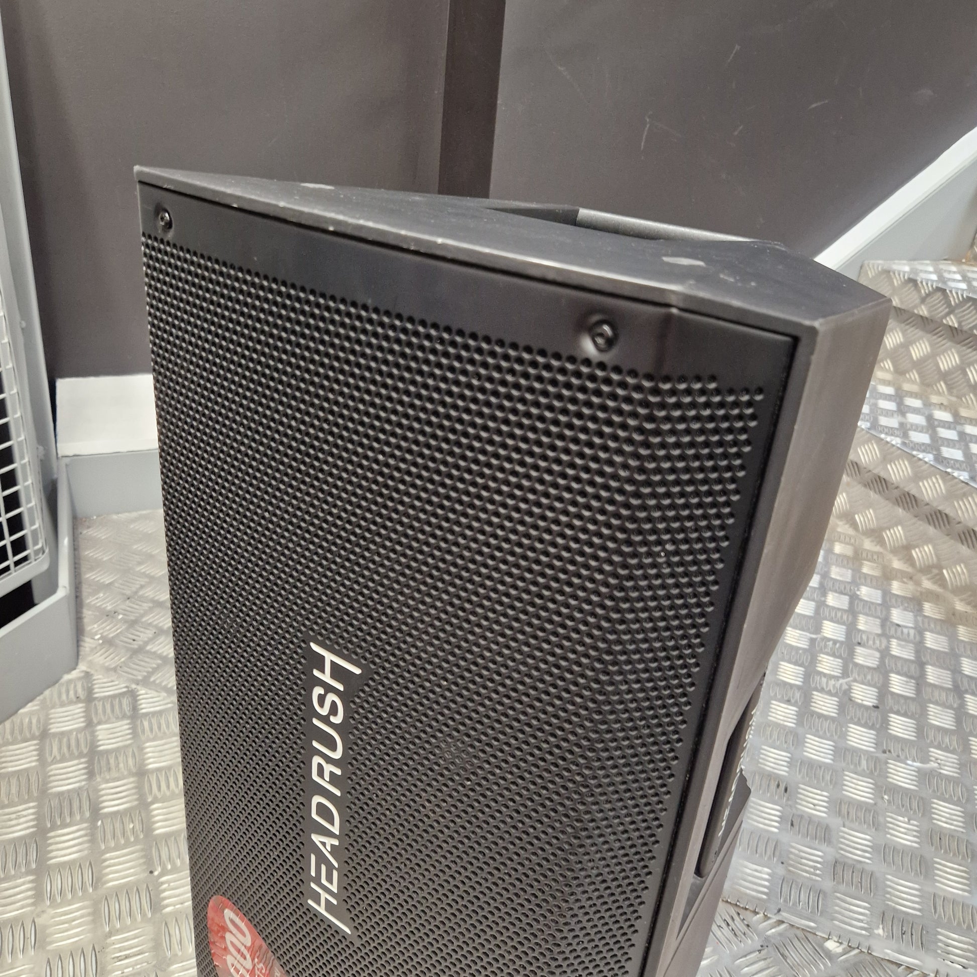 Pre-Owned Headrush FRFR-112 Full Range Powered 1x12 Speaker A black and gray portable speaker with a metal mesh grille, featuring the brand name 'HEAD RUSH' printed vertically on the front. The speaker is photographed on a diamond plate metal floor against a dark gray background.