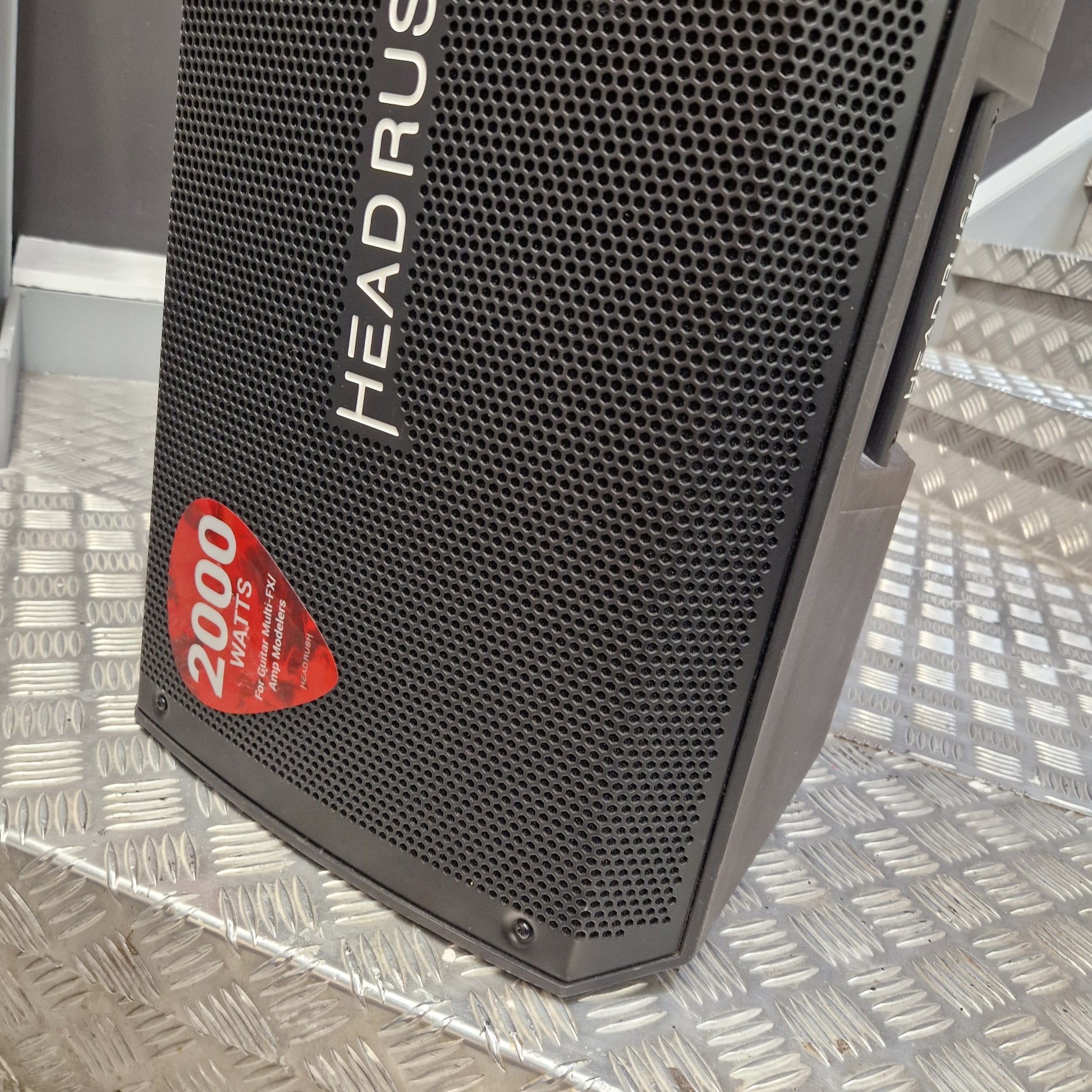Pre-Owned Headrush FRFR-112 Full Range Powered 1x12 Speaker A close-up of a black HEADRUS speaker with a red 2000 WAC label, resting on a silver diamond plate metal surface.