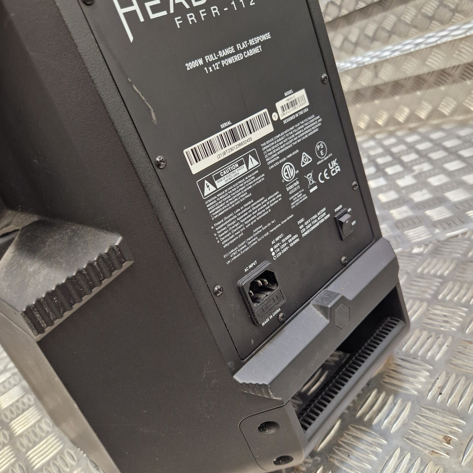 Pre-Owned Headrush FRFR-112 Full Range Powered 1x12 Speaker Close-up of the rear panel of a black HEBEY FRFR-112 powered speaker, showing the power input, barcode, and regulatory labels, resting on a diamond plate metal surface.