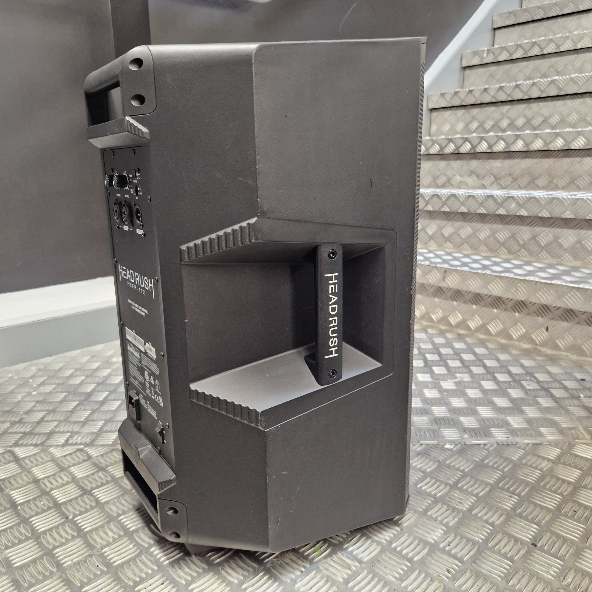 Pre-Owned Headrush FRFR-112 Full Range Powered 1x12 Speaker A dark gray Headrush portable speaker shown from a three-quarter angle, standing on a diamond plate metal floor with metal stairs in the background.