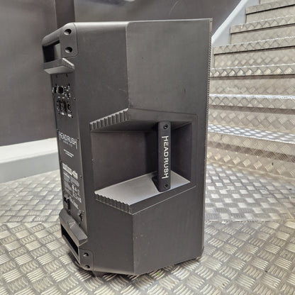 Pre-Owned Headrush FRFR-112 Full Range Powered 1x12 Speaker A dark gray Headrush portable speaker shown from a three-quarter angle, standing on a diamond plate metal floor with metal stairs in the background.