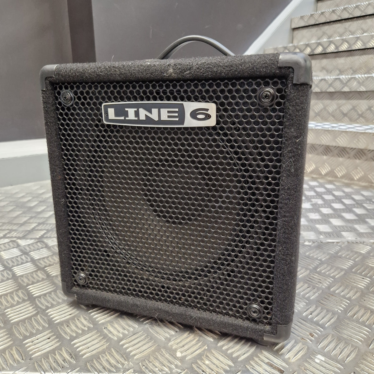 Pre-Owned Line 6 LowDown Studio 110 75w Digital Bass Combo A black Line 6 guitar amplifier with a front mesh grille and a visible 'LINE 6' logo plate, photographed on a metal diamond-plate surface with stairs in the background.