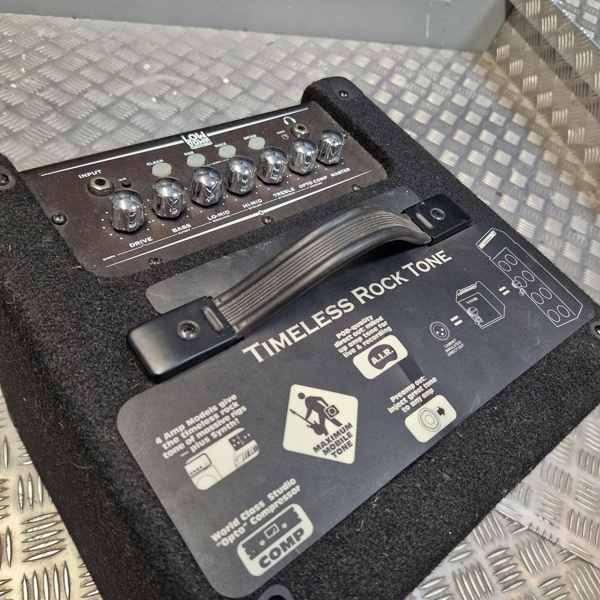 Pre-Owned Line 6 LowDown Studio 110 75w Digital Bass Combo A close-up of a black Fender Timeless Rock Tone portable guitar amp head resting on a diamond plate metal surface. The front features a black handle, a black label with white text and icons, and a dark control panel with silver knobs at the top.
