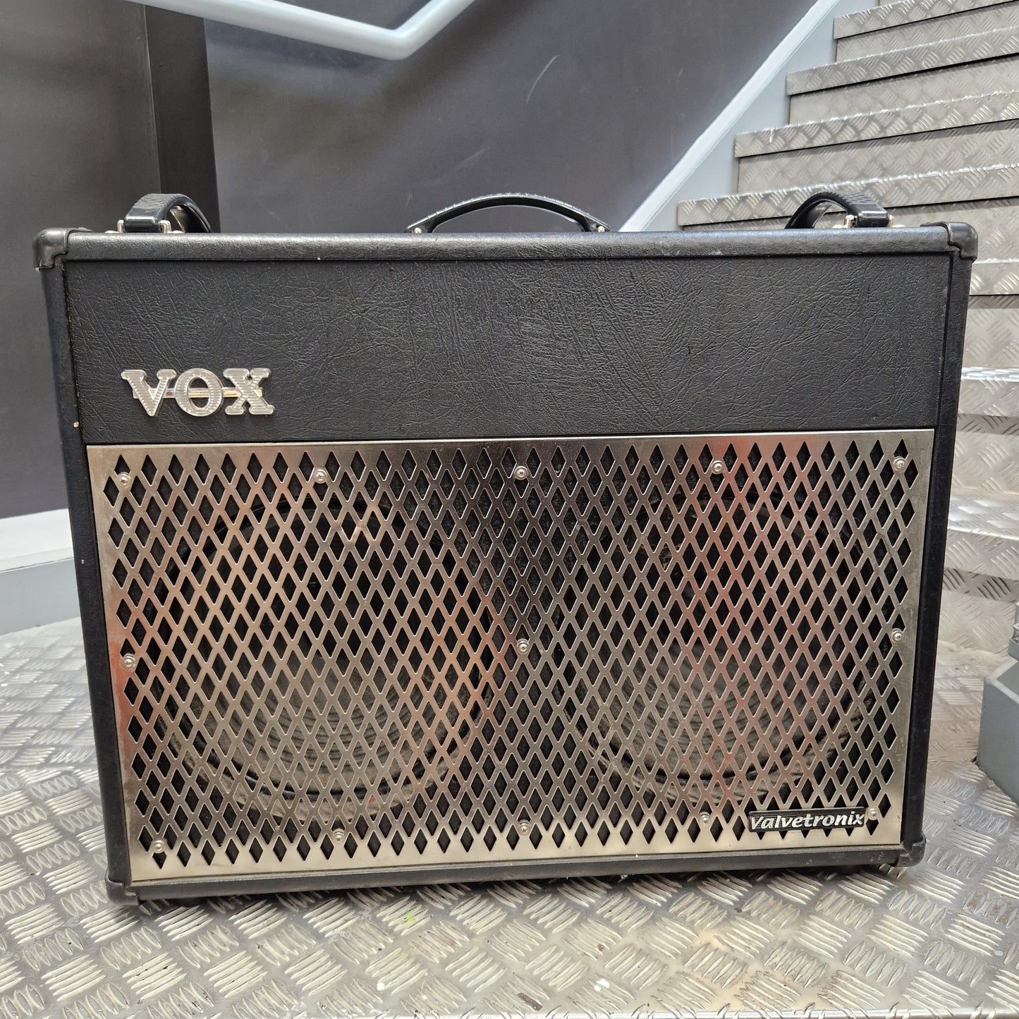 Pre-Owned Vox VT100 Valvetronix 100w Electric Combo A Vox Hemicloud 100W guitar amplifier with a black textured cabinet, silver metal grille, and silver 'VOX' logo on the front, photographed on a diamond plate metal floor in front of a staircase.