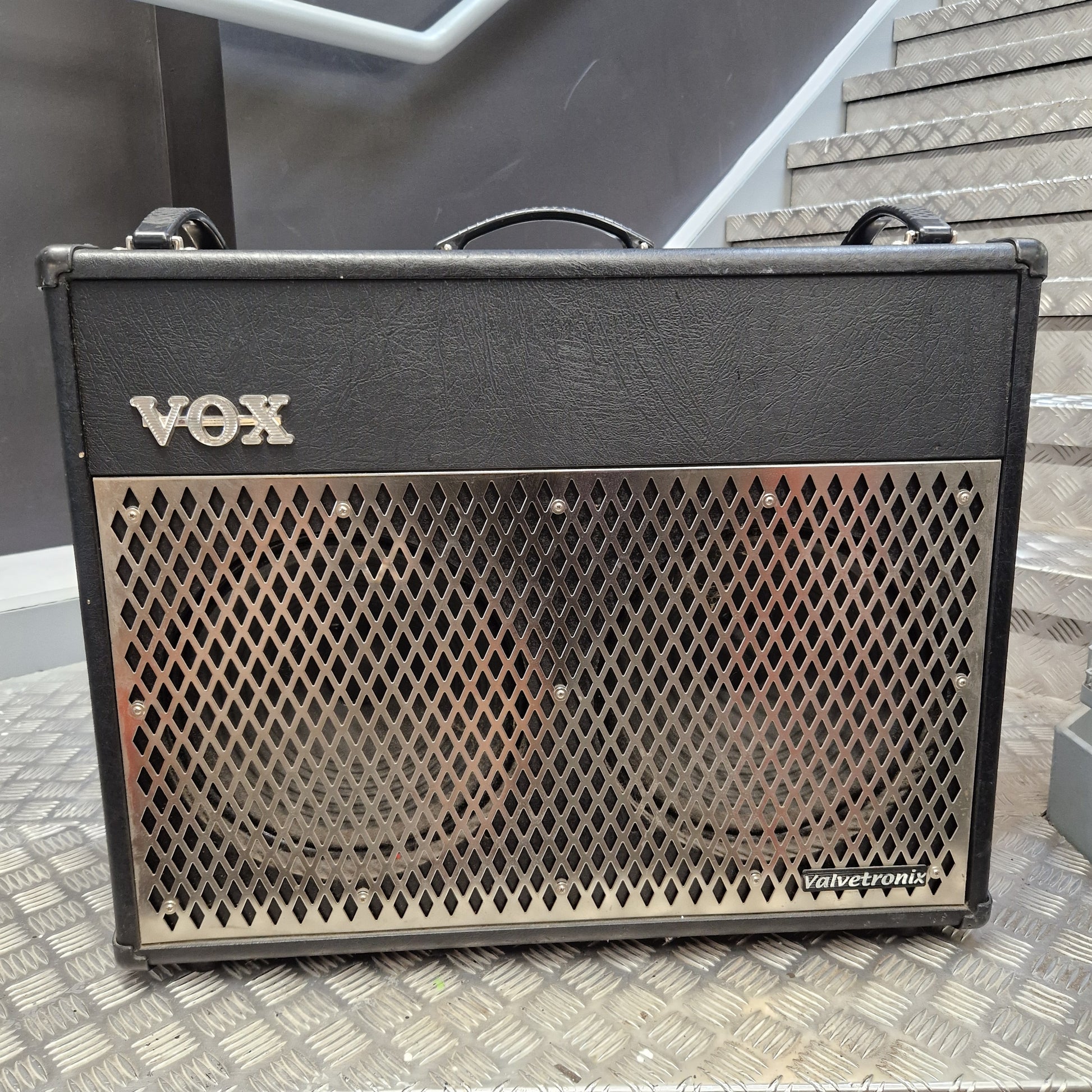 Pre-Owned Vox VT100 Valvetronix 100w Electric Combo A Vox Hemicloud 100W guitar amplifier with a black textured cabinet, silver metal grille, and silver 'VOX' logo on the front, photographed on a diamond plate metal floor in front of a staircase.