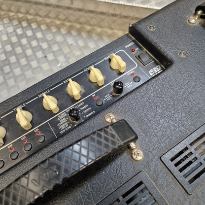 Pre-Owned Vox VT100 Valvetronix 100w Electric Combo Close-up of a black guitar amplifier control panel with cream-colored knobs, a black foot pedal, and a textured black cabinet on a diamond plate floor.