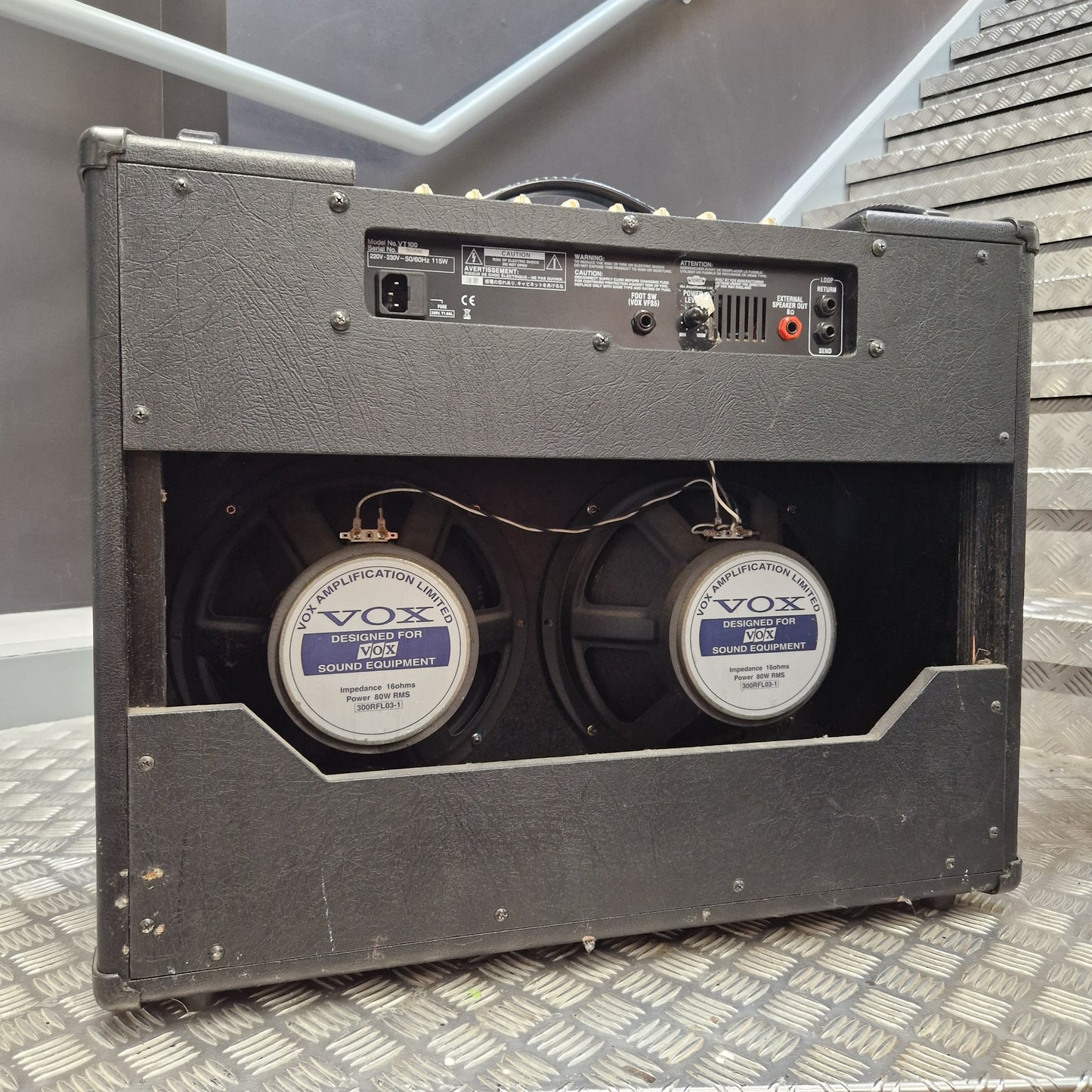 Pre-Owned Vox VT100 Valvetronix 100w Electric Combo A black Vox double speaker guitar amplifier shown from the rear, featuring two large speakers with Vox Registration 8000 labels, set against a gray staircase background.