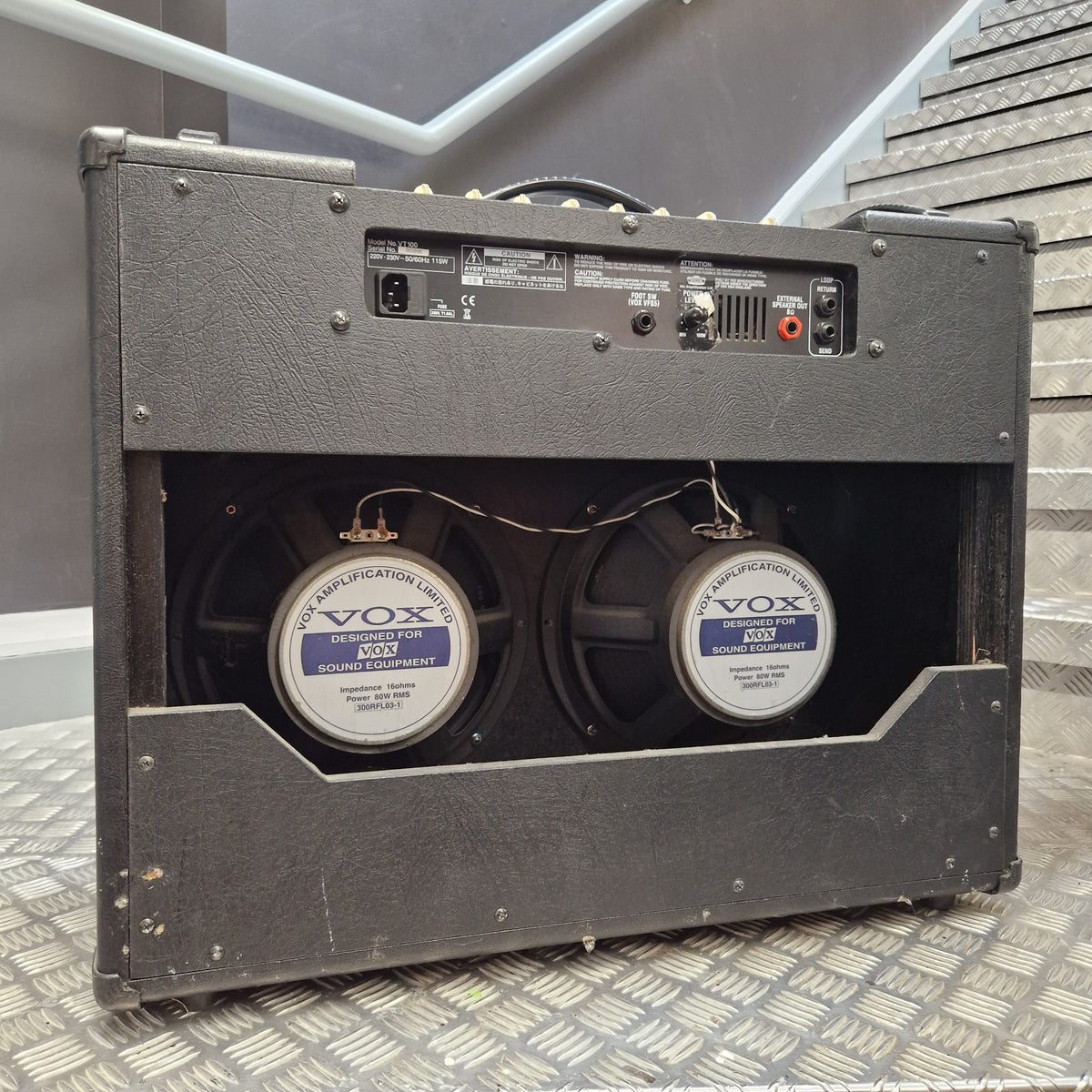 Pre-Owned Vox VT100 Valvetronix 100w Electric Combo A black Vox double speaker guitar amplifier shown from the rear, featuring two large speakers with Vox Registration 8000 labels, set against a gray staircase background.