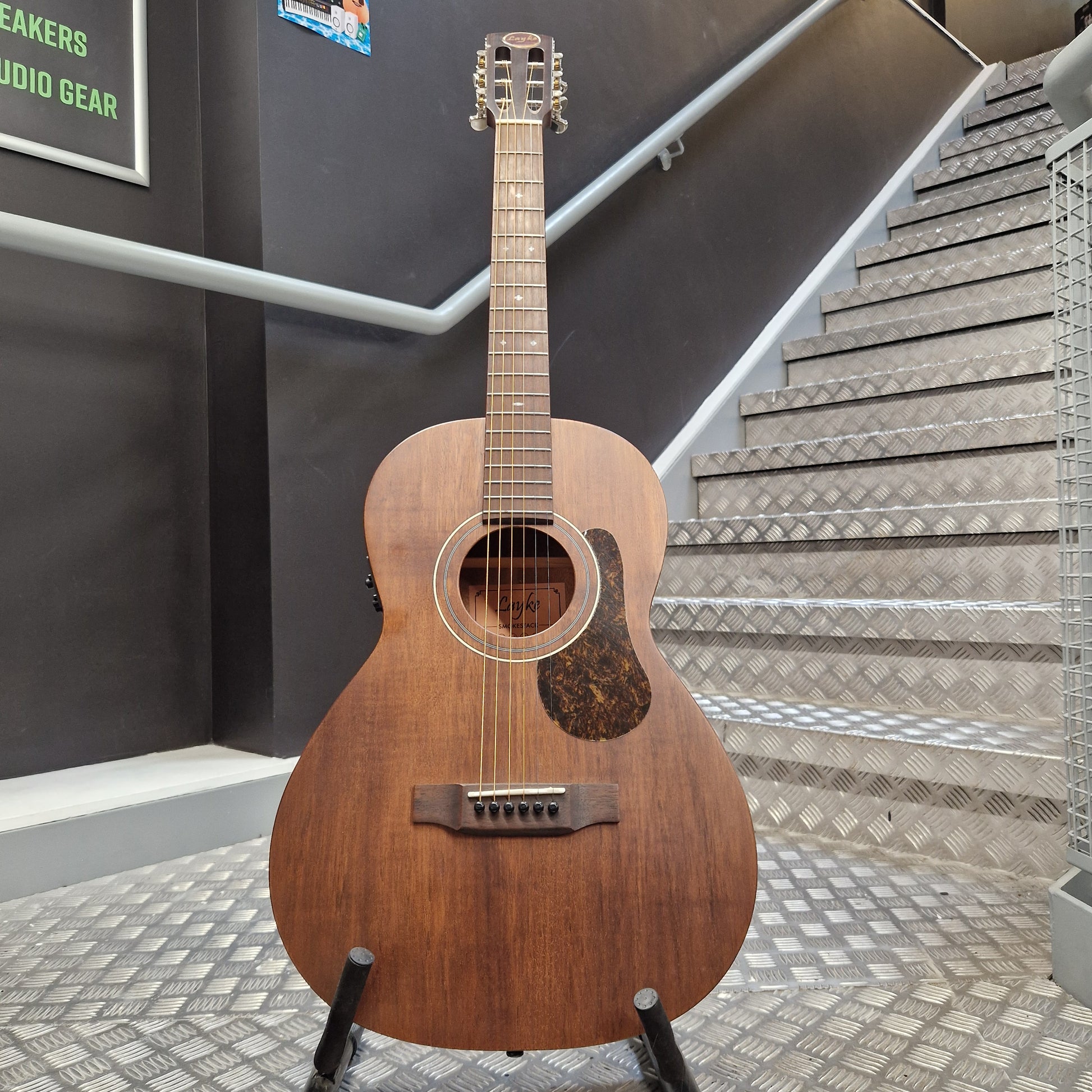 Pre-Owned Layke Smokestack Parlour Electro-Acoustic - Mahogany A vintage acoustic guitar with a medium brown wooden body and a dark tortoiseshell pickguard, displayed on a stand in front of a staircase.