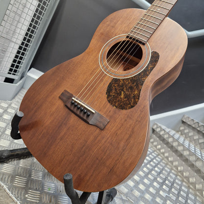 Pre-Owned Layke Smokestack Parlour Electro-Acoustic - Mahogany A wooden acoustic guitar with a medium brown finish and a dark tortoiseshell-style pickguard, shown resting on a black stand in a workshop setting.