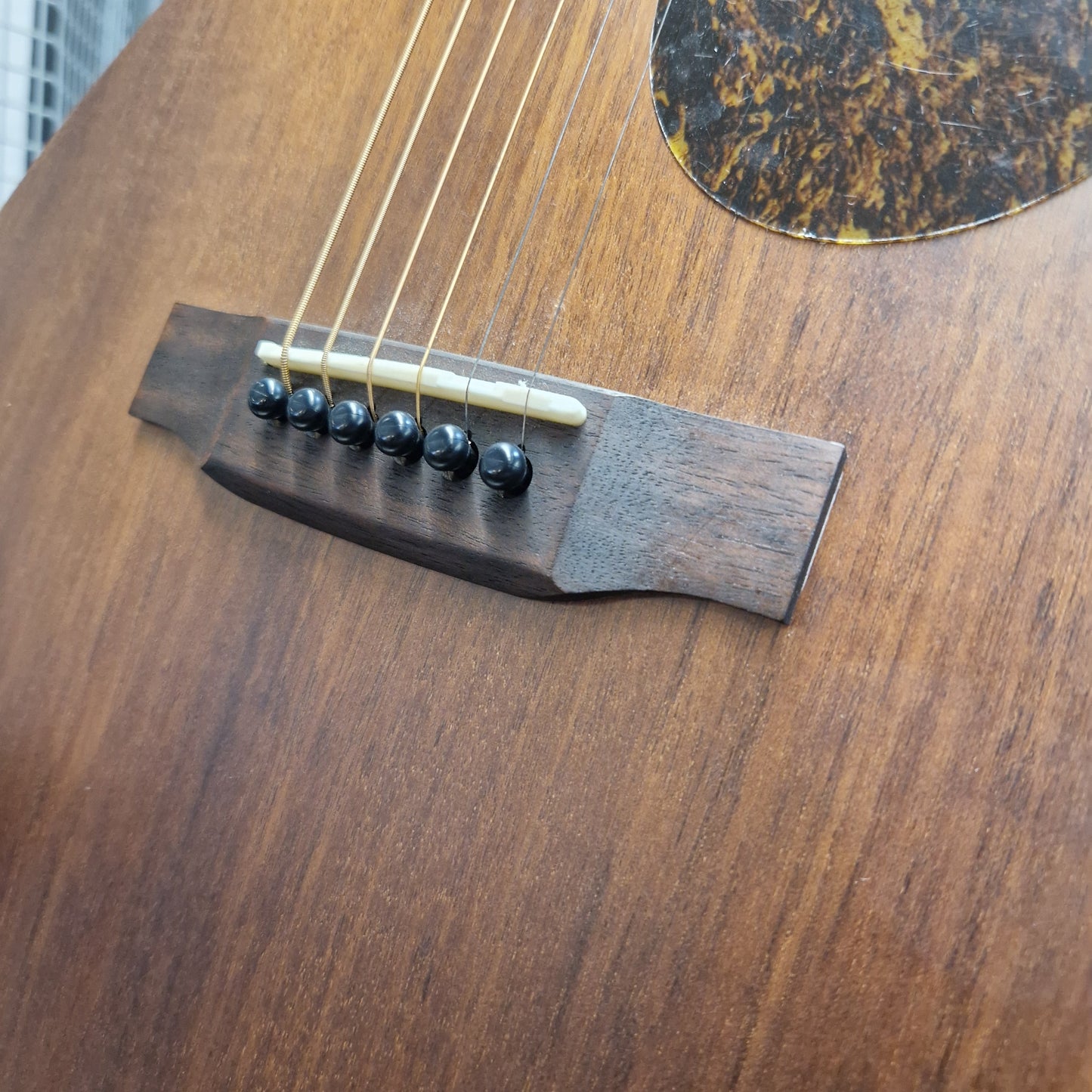 Pre-Owned Layke Smokestack Parlour Electro-Acoustic - Mahogany Close-up of a guitar's wooden bridge with six black saddle-shaped pins holding strings, set against a warm brown wood body.