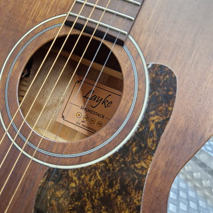 Pre-Owned Layke Smokestack Parlour Electro-Acoustic - Mahogany Close-up of a wooden acoustic guitar showing the sound hole, strings, brand label reading Layke Smokestack, and a dark tortoiseshell-style pickguard.