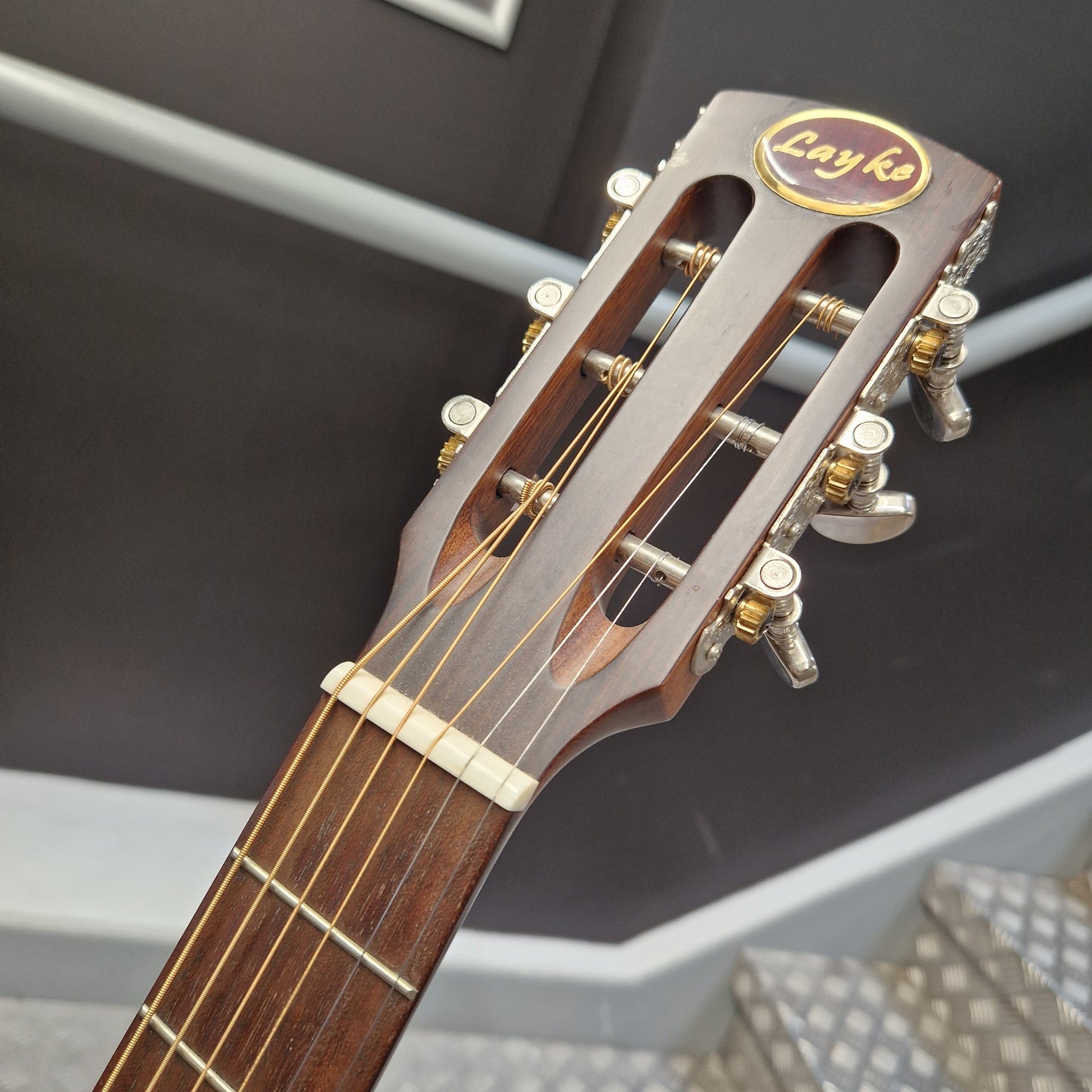 Pre-Owned Layke Smokestack Parlour Electro-Acoustic - Mahogany Close-up of the headstock of a classical guitar, showing the Layka brand logo, tuning pegs, and strings against a dark indoor background.