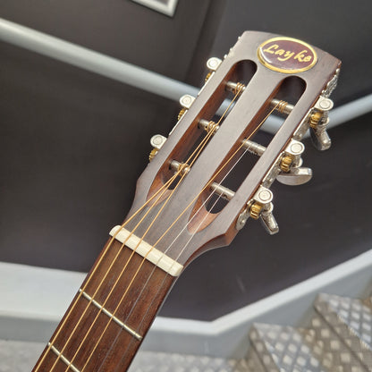 Pre-Owned Layke Smokestack Parlour Electro-Acoustic - Mahogany Close-up of the headstock of a classical guitar, showing the Layka brand logo, tuning pegs, and strings against a dark indoor background.