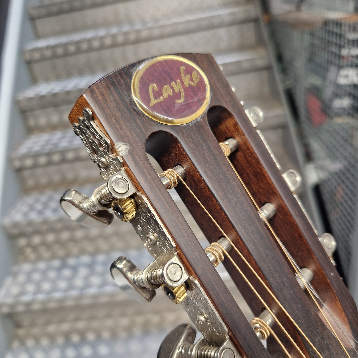 Pre-Owned Layke Smokestack Parlour Electro-Acoustic - Mahogany Close-up of a wooden Layke guitar headstock with gold tuning pegs and strings, set against a blurred staircase background.