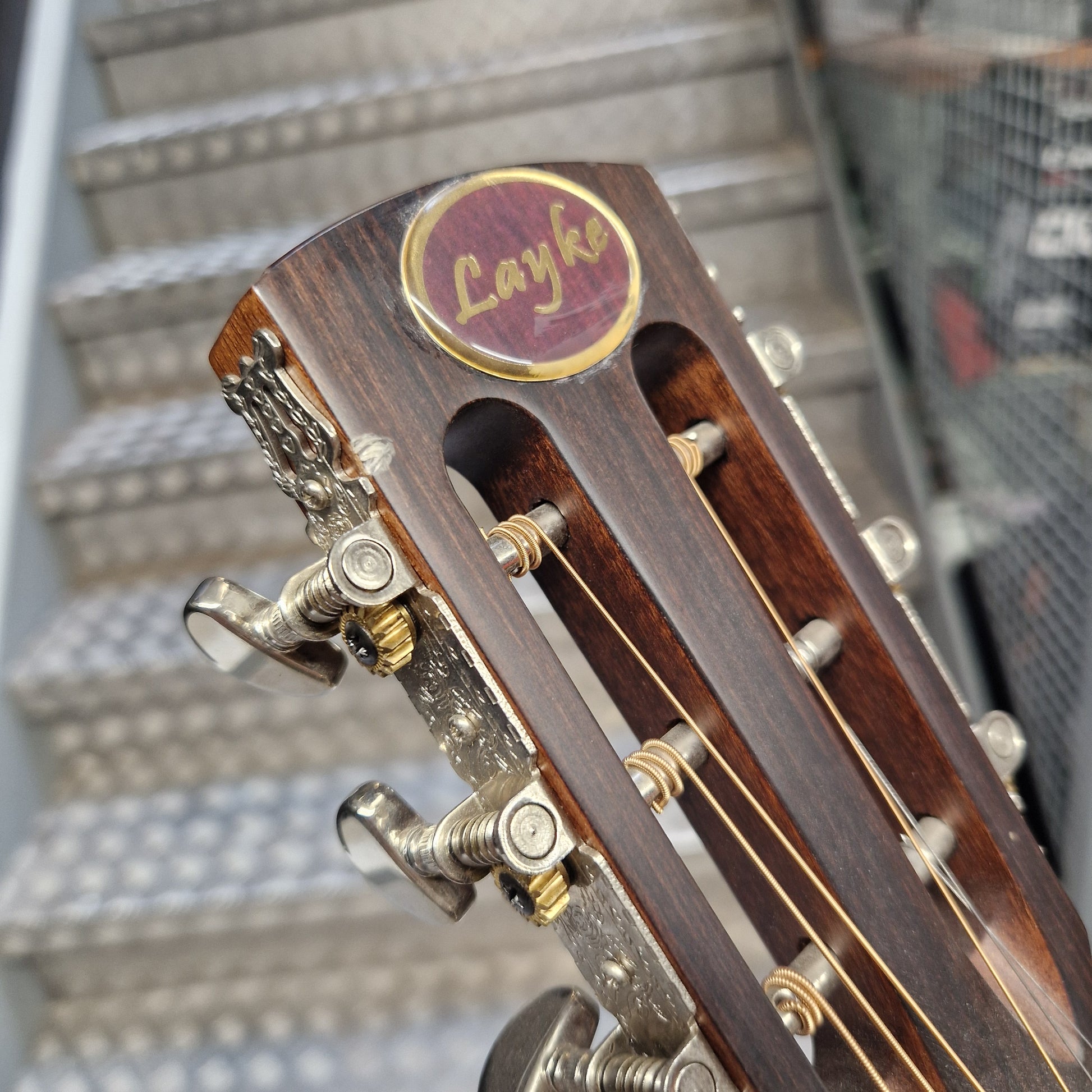 Pre-Owned Layke Smokestack Parlour Electro-Acoustic - Mahogany Close-up of a wooden Layke guitar headstock with gold tuning pegs and strings, set against a blurred staircase background.