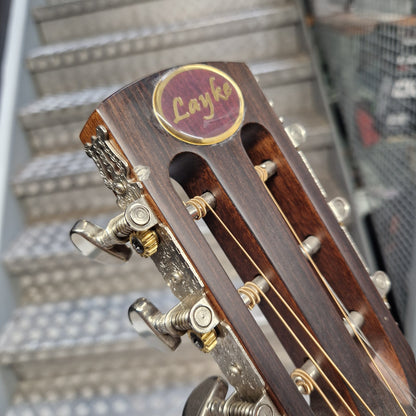 Pre-Owned Layke Smokestack Parlour Electro-Acoustic - Mahogany Close-up of a wooden Layke guitar headstock with gold tuning pegs and strings, set against a blurred staircase background.