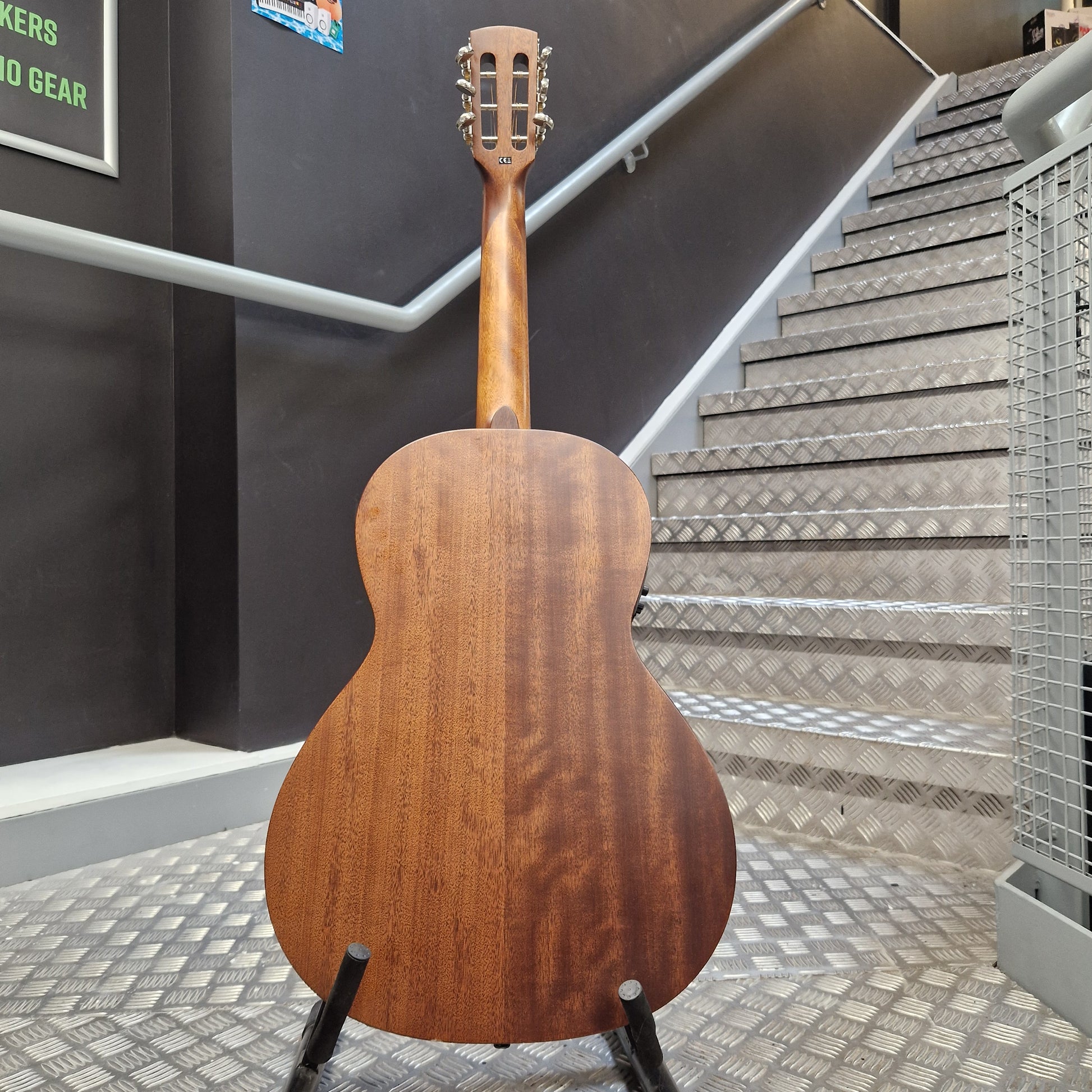 Pre-Owned Layke Smokestack Parlour Electro-Acoustic - Mahogany A wooden acoustic guitar shown from the back, resting on a black stand in an indoor setting with a staircase and dark gray walls in the background.