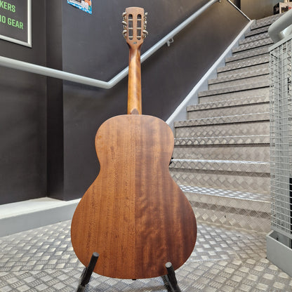 Pre-Owned Layke Smokestack Parlour Electro-Acoustic - Mahogany A wooden acoustic guitar shown from the back, resting on a black stand in an indoor setting with a staircase and dark gray walls in the background.