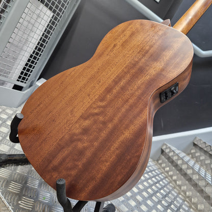 Pre-Owned Layke Smokestack Parlour Electro-Acoustic - Mahogany Close-up of the back of a wooden acoustic guitar on a stand, showing the rich brown wood grain finish and a black output jack, set against a workshop background with metal steps.