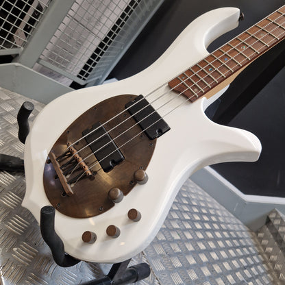 Pre-Owned Traben Neo 4 Bass - White A white electric bass guitar with a distinctive oval brown pickguard, mounted on a black stand in an industrial setting with a diamond plate floor.