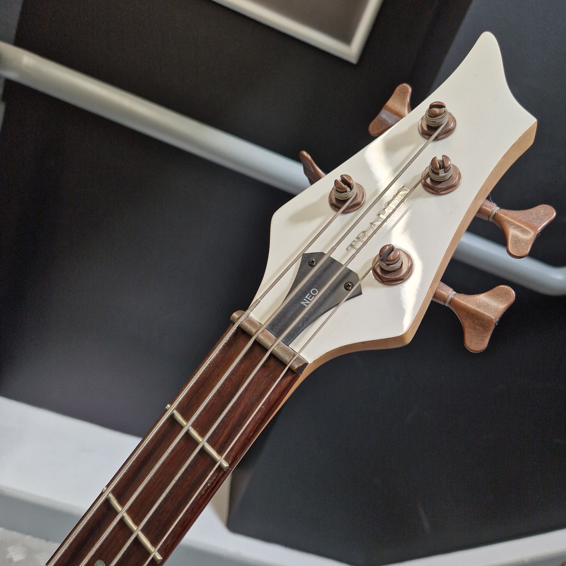 Pre-Owned Traben Neo 4 Bass - White Close-up of a bass guitar headstock and neck, showing a white glossy headstock with four tuning pegs, a dark brown fretboard, and strings. The brand name 'NEO' is visible on the headstock.