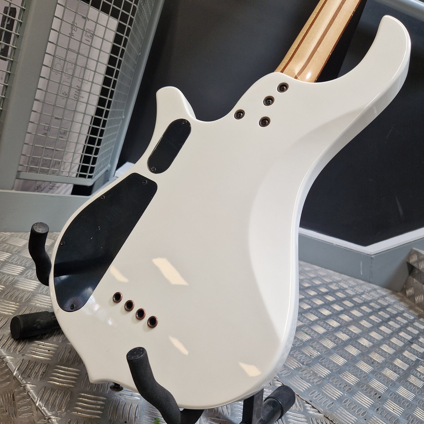 Pre-Owned Traben Neo 4 Bass - White Close-up of the back of a white electric guitar resting on a black stand on a metal diamond plate floor, with a dark wall and industrial background.