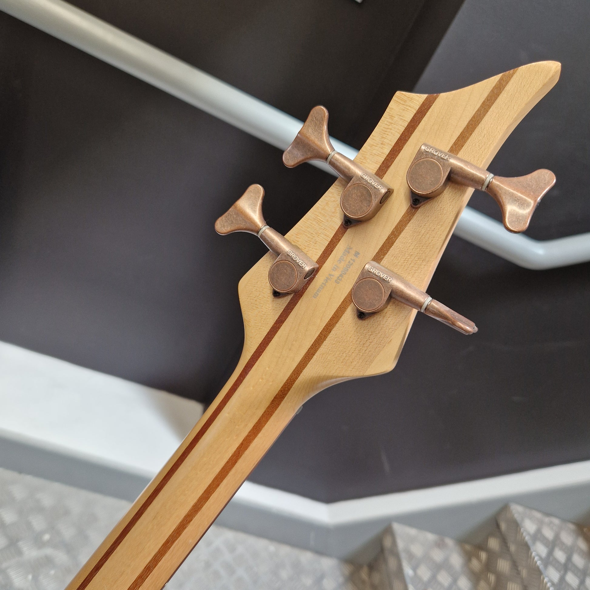 Pre-Owned Traben Neo 4 Bass - White Close-up of a bass guitar headstock with four metallic tuning pegs and wood inlays, photographed indoors against a dark wall and metal steps.