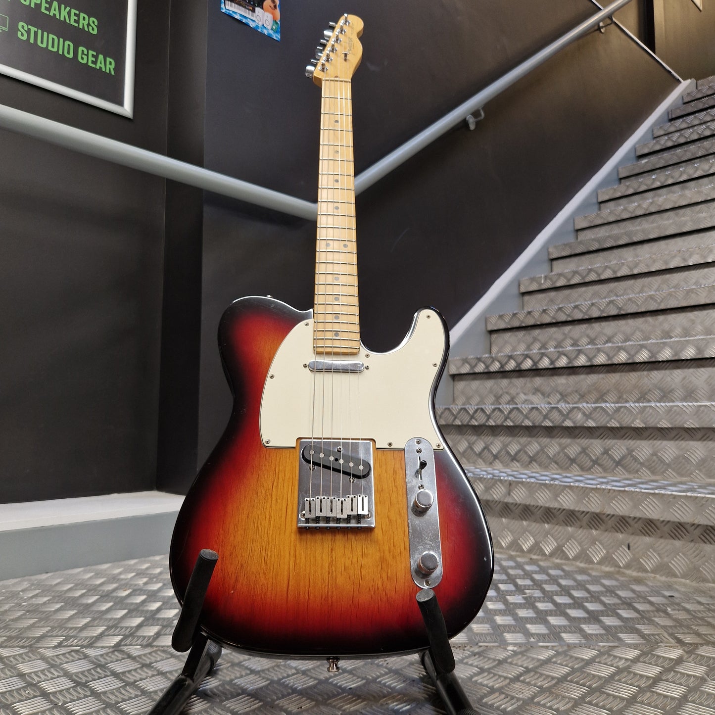 Pre-Owned Fender American Standard Telecaster 2000 Electric - 3-Colour Sunburst A classic Fender Telecaster electric guitar with a sunburst finish, standing on a black stand in a shop or studio setting with a staircase in the background.