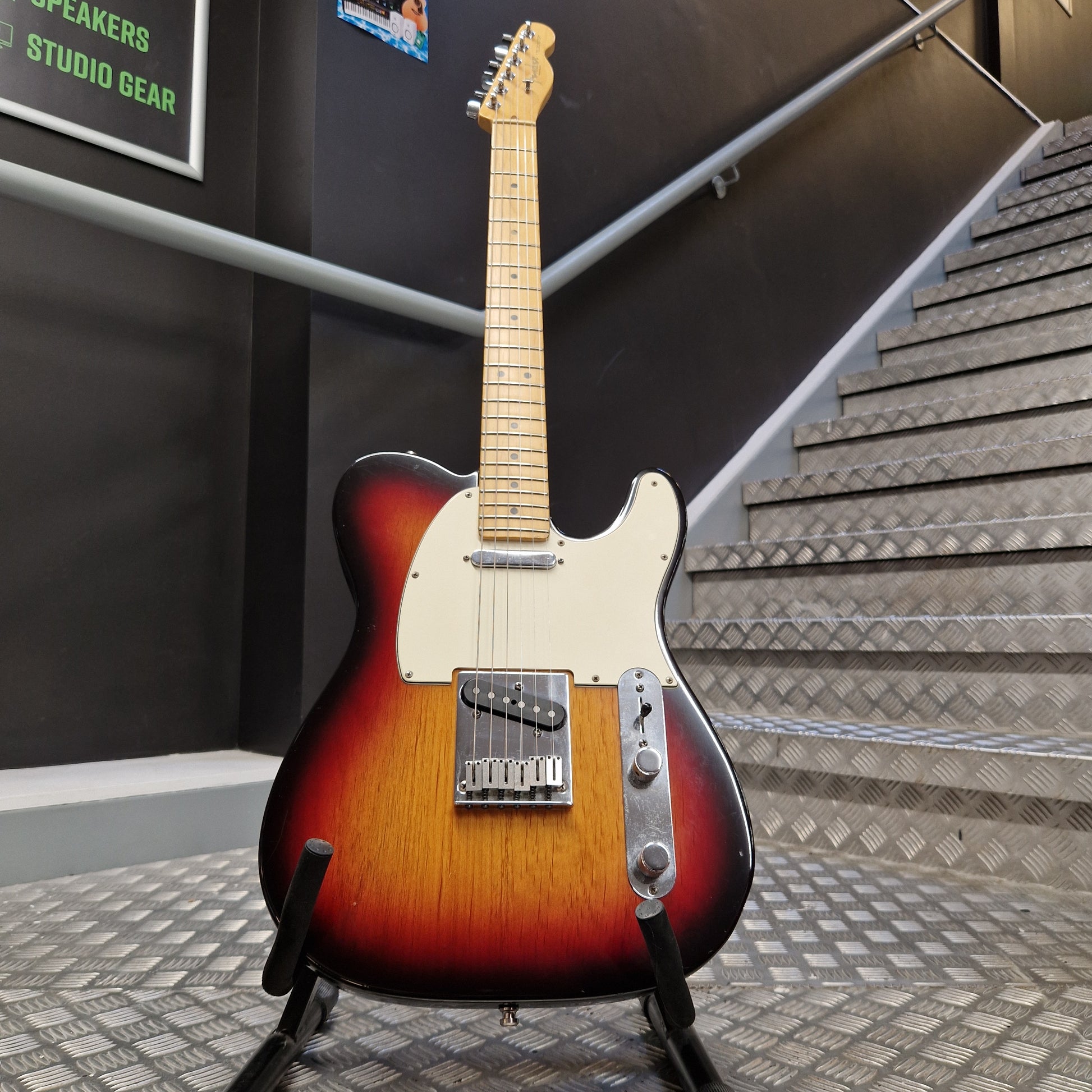 Pre-Owned Fender American Standard Telecaster 2000 Electric - 3-Colour Sunburst A classic Fender Telecaster electric guitar with a sunburst finish, standing on a black stand in a shop or studio setting with a staircase in the background.