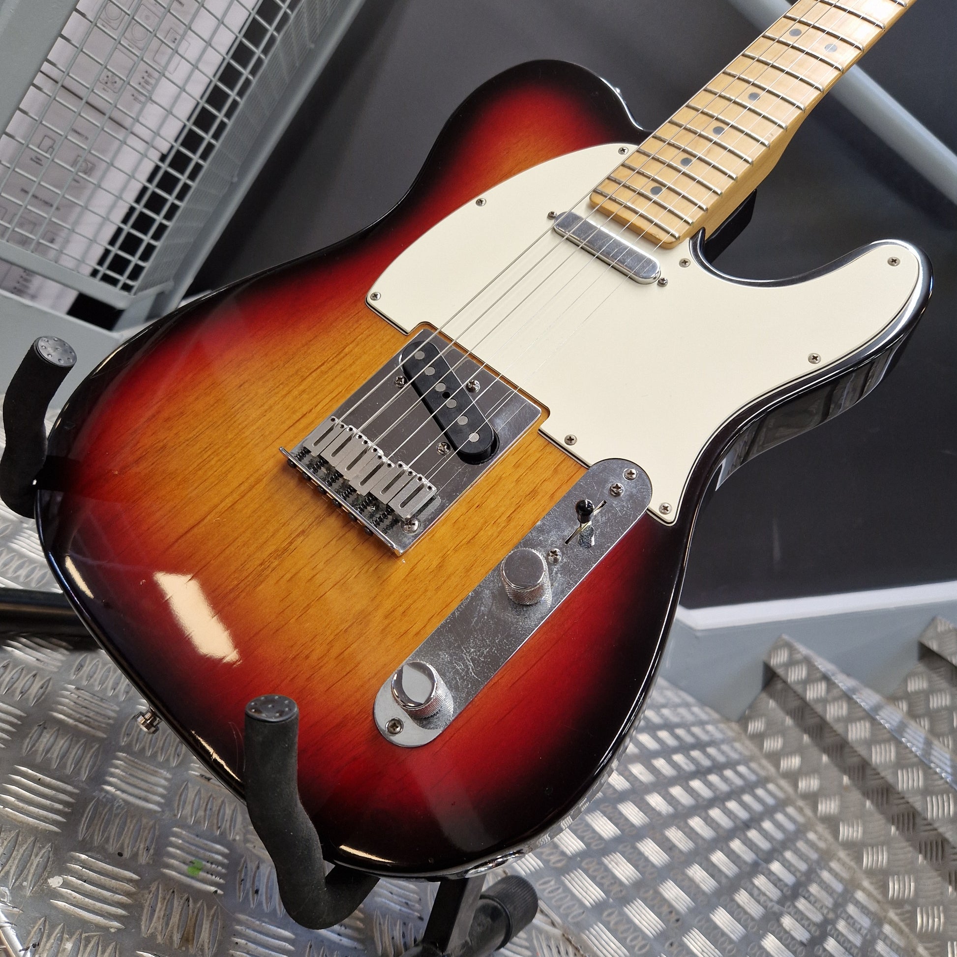 Pre-Owned Fender American Standard Telecaster 2000 Electric - 3-Colour Sunburst A classic Fender Telecaster-style electric guitar with a sunburst finish, cream pickguard, and maple neck, resting on a black stand over a textured metal surface.