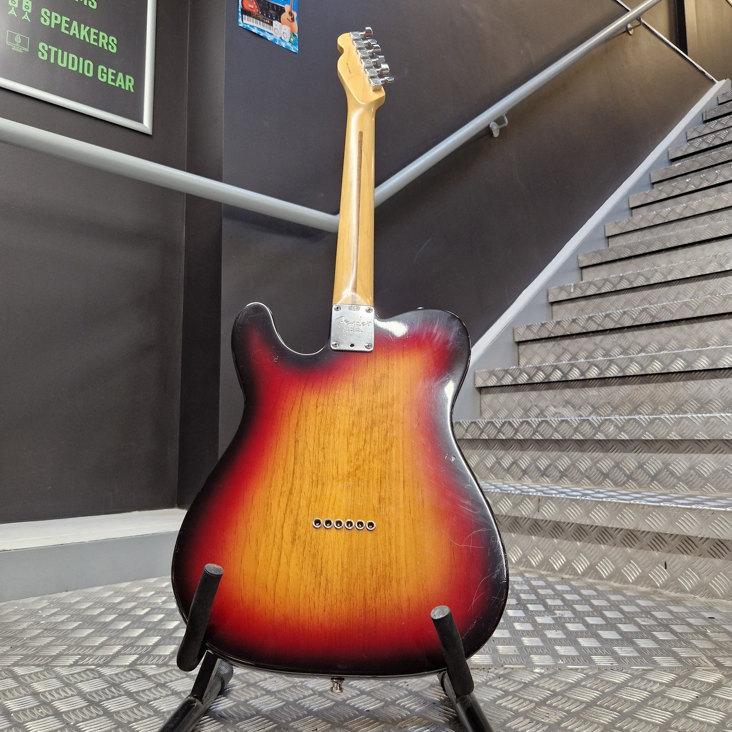 Pre-Owned Fender American Standard Telecaster 2000 Electric - 3-Colour Sunburst A classic electric guitar with a sunburst finish, standing on a black stand in an indoor setting with a staircase and dark gray walls in the background.