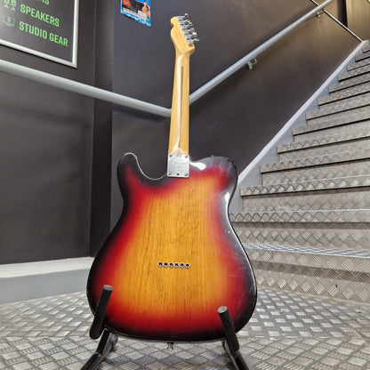 Pre-Owned Fender American Standard Telecaster 2000 Electric - 3-Colour Sunburst A classic electric guitar with a sunburst finish, standing on a black stand in an indoor setting with a staircase and dark gray walls in the background.