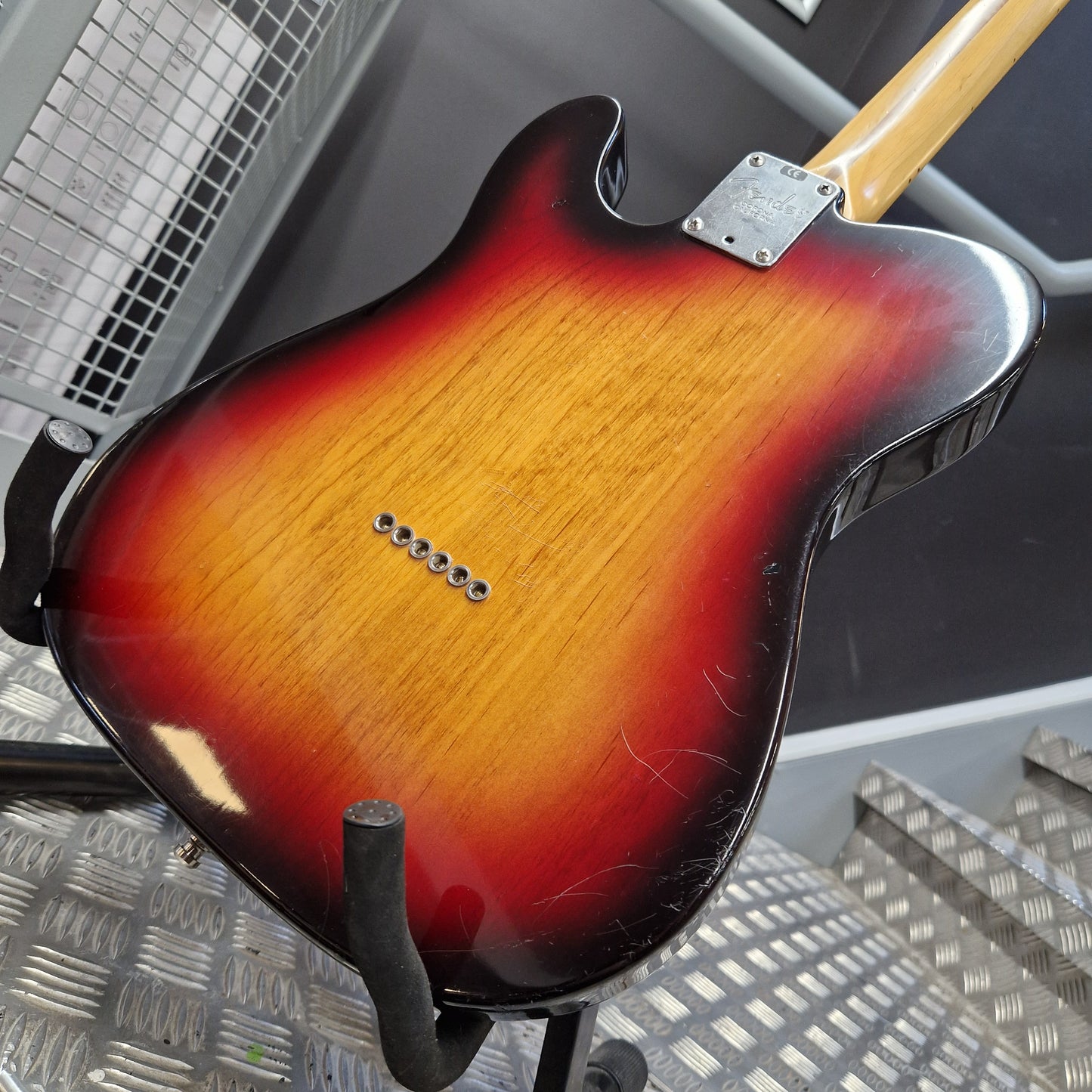 Pre-Owned Fender American Standard Telecaster 2000 Electric - 3-Colour Sunburst Close-up of the back of a double-cutaway electric guitar with a sunburst finish, resting on a stand in a workshop setting.