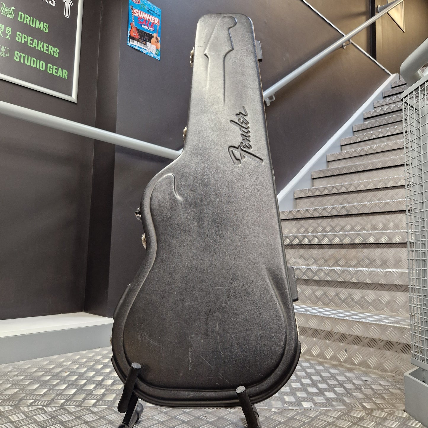 Pre-Owned Fender American Standard Telecaster 2000 Electric - 3-Colour Sunburst A used black Fender guitar hard case standing on a small display stand in an indoor setting with a metal staircase and dark gray walls in the background.