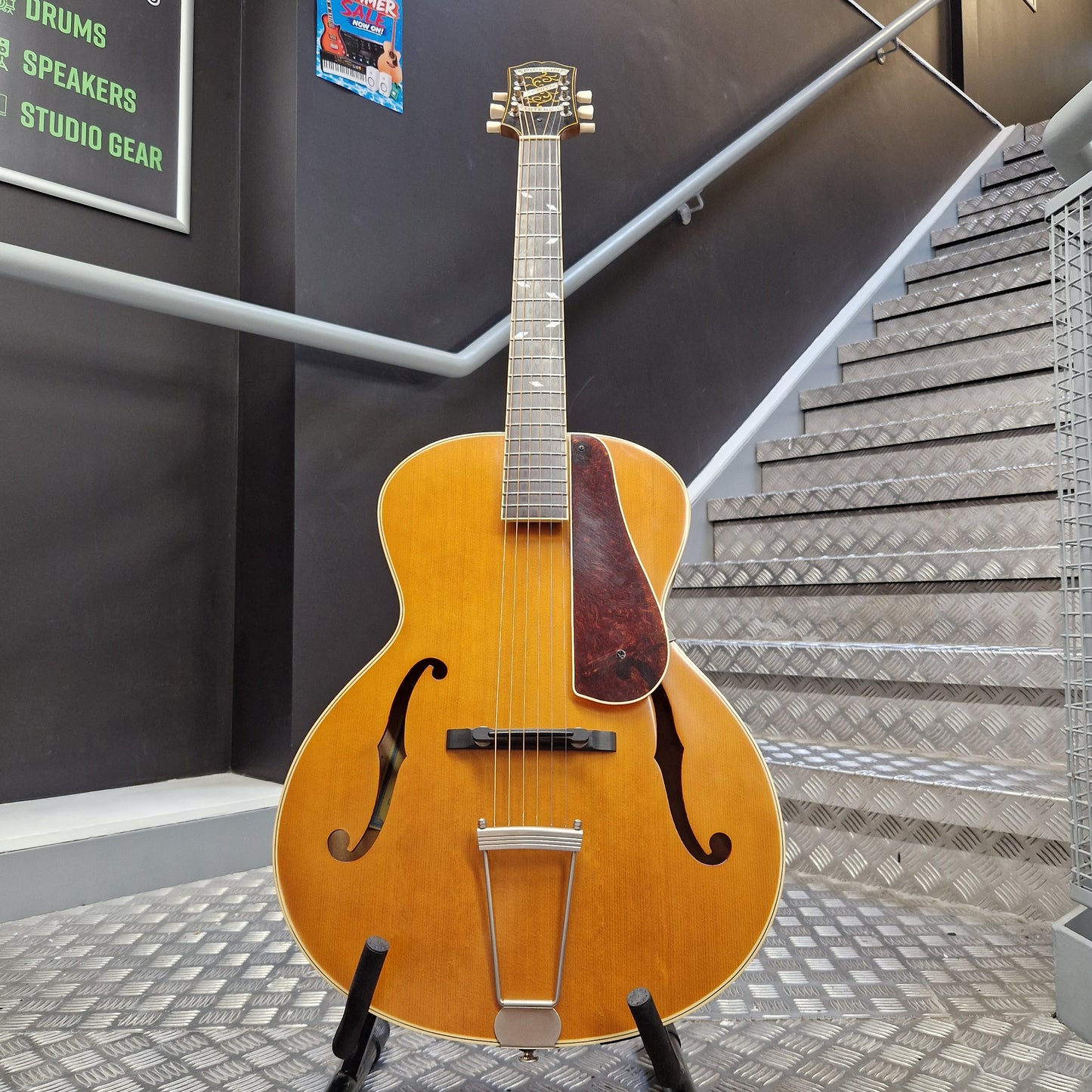 Pre-Owned Epiphone Masterbilt Zenith Classic Electro-Acoustic - Vintage Natural A vintage-style hollow-body electric guitar with a natural wood finish stands on a black stand in front of a staircase and dark wall.